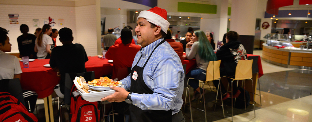 A UIW employee wearing a Santa hat carries a bowl of food