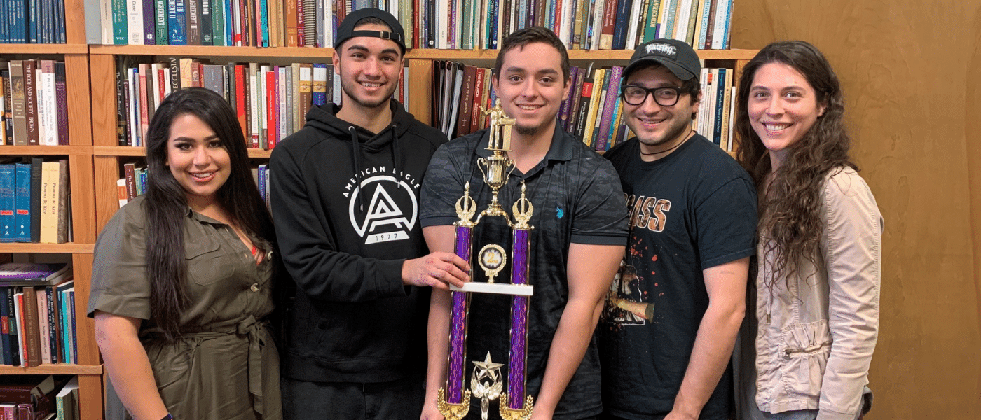 Four winners of the Ethics Bowl holding a large trophy in front of a bookshelves.