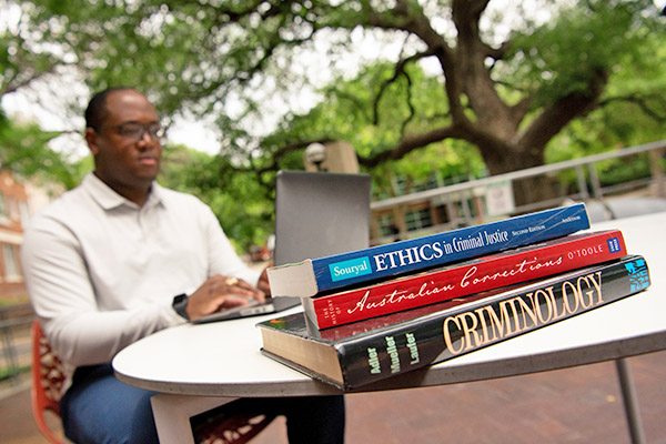 Close up of criminal justice books on an outdoor table with a student typing on his laptop in the backg
