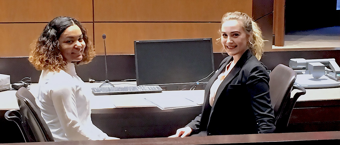 Two students smiling at the camera while sitting at the legal council table in a court