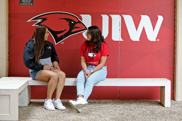 Current UIW students sitting on a bench in front of the UIW cardinal logo.