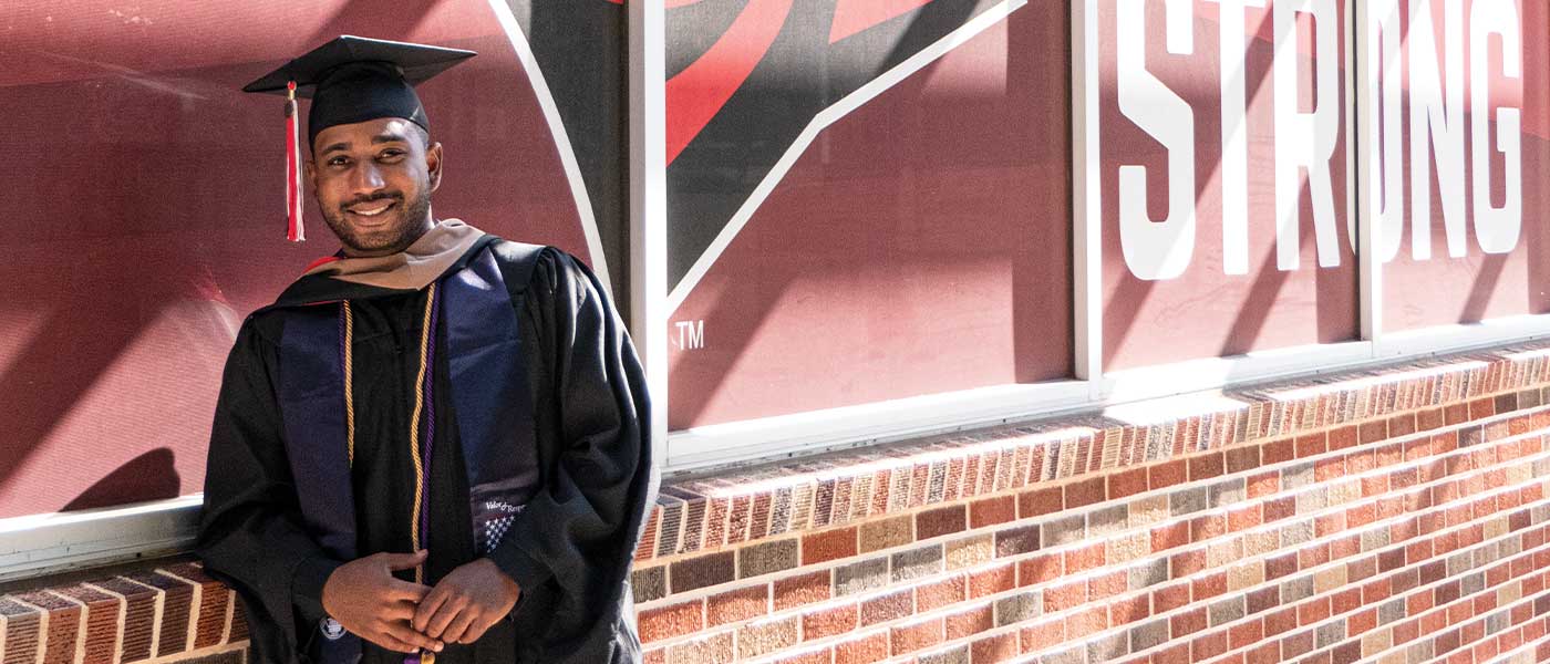 UIW veteran student in cap and graduation gown in front of the flags and chapel 