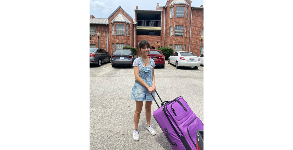 Student poses with a suitcase in front of an apartment building